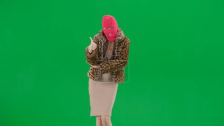 Foto de Mujer en pasamontañas rosa, abrigo de tigre y vestido de noche muestra el gesto de los pulgares hacia arriba. Una mujer loca sobre un fondo verde en un estudio. Concepto de tendencia de moda, tendencia feminista en la moda - Imagen libre de derechos