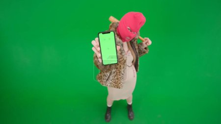 Foto de Mujer en pasamontañas rosa, abrigo de tigre y vestido mostrando el teléfono de pantalla verde a la cámara. Mujer loca con bate en las manos sobre fondo verde en el estudio. Concepto de tendencia de moda, tendencia feminista en la moda - Imagen libre de derechos