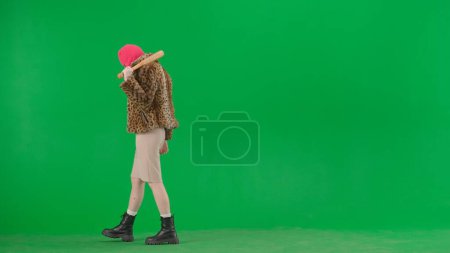 Téléchargez les photos : Femme en cagoule rose, manteau de tigre, robe de soirée et avec une batte dans les mains. Femme bizarre sur fond vert en studio. Concept de tendance mode - en image libre de droit