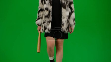 Photo for Woman in fur coat and evening dress walking with bat in hand. Woman freak on green background in studio. Fashion trend concept, feminist trend in fashion - Royalty Free Image
