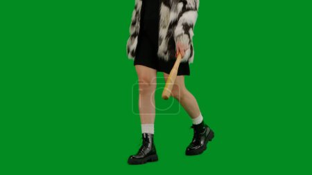 Photo for Woman in fur coat and evening dress walking with bat in hand. Woman freak on green background in studio. Fashion trend concept, feminist trend in fashion - Royalty Free Image
