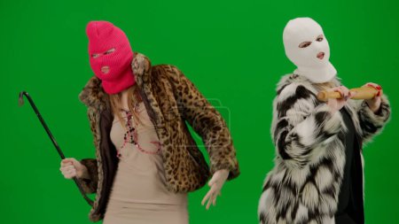 Photo for Two women in white and pink balaclavas dancing funny with a bat and a crowbar. Freak women in fur coats on green studio background. Fashion trend concept, feminist trend in fashion - Royalty Free Image