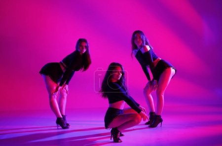 Téléchargez les photos : Groupe de trois femmes posant dans un studio en lumière rose fluo. Danseurs en short noir et hauts courts et talons hauts. Chorégraphie contemporaine. Sur toute la longueur. Promo ou commercial - en image libre de droit