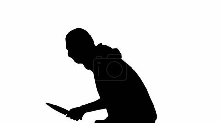 Foto de Silueta negra de ladrón sobre fondo blanco aislado. Un ladrón con capucha y pasamontañas camina con un cuchillo en las manos, preparándose para cometer un crimen. Vista lateral - Imagen libre de derechos