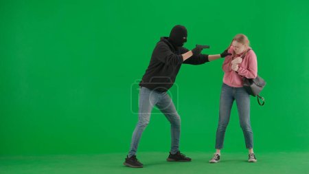 Photo for Robbery and crime advertising concept. Portrait of thief and victim against green screen with chromakey. Male robber in balaclava with a gun attacks a girl - Royalty Free Image