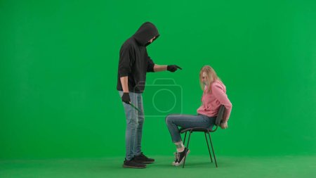 Photo for Robbery and criminal concept. Portrait of thief and victim on chroma key green screen background. Girl sitting on chair tied hands and taped mouth man robber with knife threatens her. - Royalty Free Image