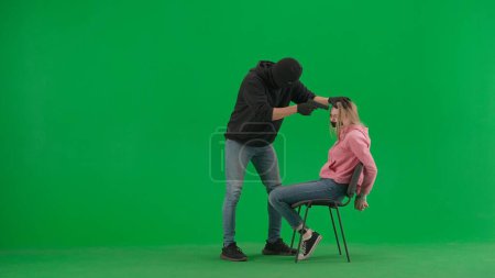 Photo for Robbery and criminal concept. Portrait of thief and victim on chroma key green screen background. Girl sitting on chair tied hands and taped mouth man robber with gun threatens her. - Royalty Free Image