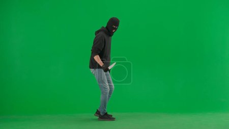 Photo for Robbery and criminal concept. Portrait of thief on chroma key green screen background. Man robber in black balaclava and hoodie holding knife looks around. - Royalty Free Image