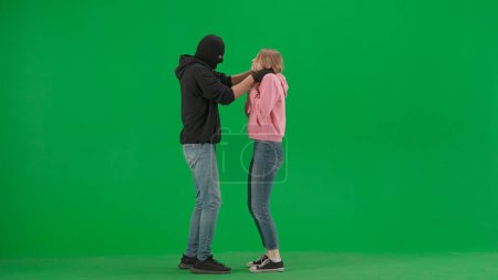 Photo for Robbery and crime advertising concept. Portrait of thief and victim on chromakey background of green screen. Male robber in black balaclava mask strangling a girl - Royalty Free Image