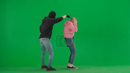 Photo for Robbery and crime advertising concept. Portrait of thief and victim on chromakey background of green screen. Male robber in black balaclava with a gun attacks a girl - Royalty Free Image