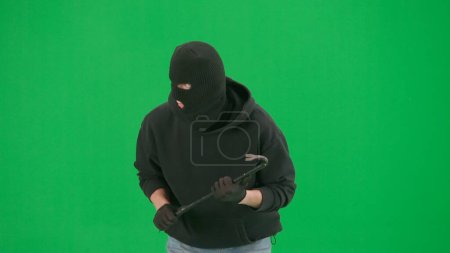 Photo for Robbery and criminal concept. Portrait of thief on chroma key green screen background. Man robber in balaclava and hoodie walking holding crowbar in hands looks around. - Royalty Free Image