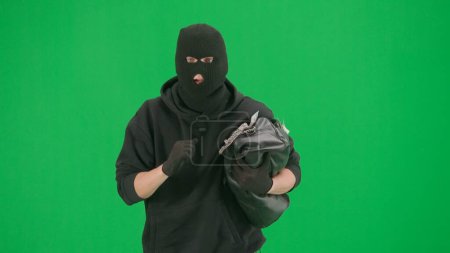 Photo for Robbery and criminal concept. Portrait of thief on chroma key green screen background. Man robber wearing hoodie, jeans and balaclava, running from police with stolen bag full of money. - Royalty Free Image