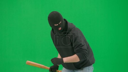 Photo for Robbery and criminal concept. Portrait of thief on chroma key green screen background. Man robber in balaclava and hoodie walking holding baseball bat in hand looks around. Half turn - Royalty Free Image