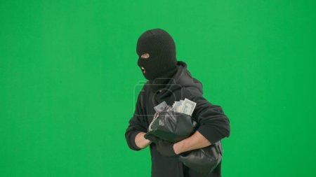 Photo for Robbery and criminal concept. Portrait of thief on chroma key green screen background. Man robber wearing hoodie, jeans and balaclava, running from police with stolen bag full of money. Side view - Royalty Free Image