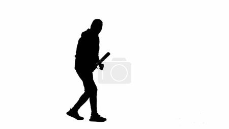 Foto de Silueta negra de ladrón sobre fondo blanco aislado. Un ladrón con capucha y pasamontañas camina con un palo de madera en las manos, preparándose para cometer un crimen. Media vuelta. - Imagen libre de derechos