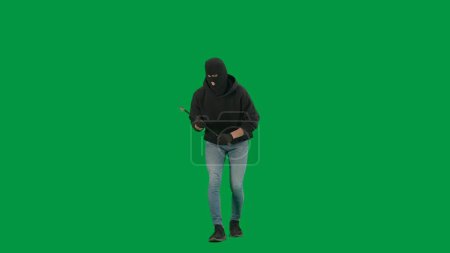 Photo for Robbery and criminal concept. Portrait of thief on chroma key green screen background. Man robber in balaclava and hoodie walking holding crowbar in hands looks around. - Royalty Free Image