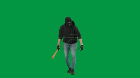 Photo for Robbery and criminal concept. Portrait of thief on chroma key green screen background. Man robber in balaclava and hoodie walking holding baseball bat in hand looks around. - Royalty Free Image