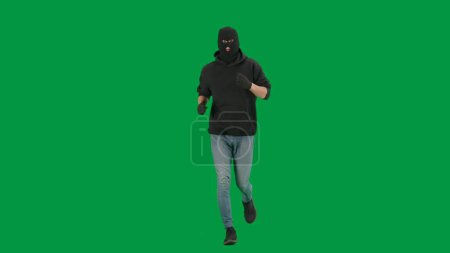 Foto de Robo y concepto criminal. Retrato de ladrón en croma pantalla verde clave de fondo. Hombre ladrón con capucha, jeans y pasamontañas negro, corriendo preparándose para cometer un crimen. - Imagen libre de derechos