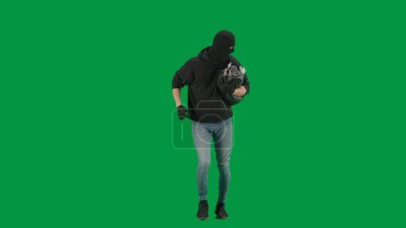 Foto de Robo y concepto criminal. Retrato de ladrón en croma pantalla verde clave de fondo. Hombre ladrón con sudadera con capucha, jeans y pasamontañas, huyendo de la policía con una bolsa robada llena de dinero. - Imagen libre de derechos