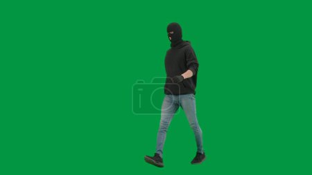 Téléchargez les photos : Vol qualifié et concept criminel. Portrait de voleur sur fond vert chroma key. Un voleur portant un sweat à capuche, un jean et de la cagoule noire, se préparant à commettre un crime. Demi-tour - en image libre de droit