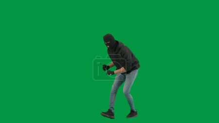Photo for Robbery and criminal concept. Portrait of thief on chroma key green screen background. Man robber in balaclava and hoodie walking holding crowbar in hands looks around. Half turn - Royalty Free Image