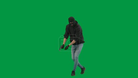 Photo for Robbery and criminal concept. Portrait of thief on chroma key green screen background. Man robber in balaclava and hoodie walking holding gun in hand looks around. Half turn - Royalty Free Image