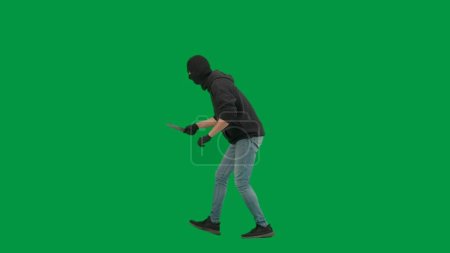 Foto de Robo y concepto criminal. Retrato de ladrón en croma pantalla verde clave de fondo. Hombre ladrón en pasamontañas y sudadera con capucha caminando con cuchillo en la mano mira a su alrededor. Vista lateral - Imagen libre de derechos