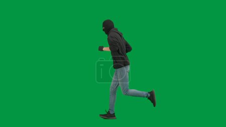 Téléchargez les photos : Vol qualifié et concept criminel. Portrait de voleur sur fond vert chroma key. Un voleur portant un sweat à capuche, un jean et de la cagoule noire, se préparant à commettre un crime. Vue latérale - en image libre de droit