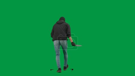 Foto de Robo y concepto criminal. Retrato de ladrón en croma pantalla verde clave de fondo. Hombre ladrón en pasamontañas y sudadera con capucha caminando con cuchillo en la mano mira a su alrededor. Vista trasera - Imagen libre de derechos