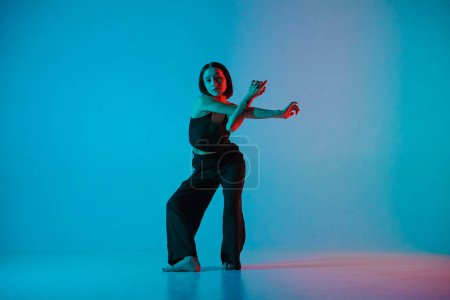 Foto de Mujer joven en pantalones negros y top posa en estudio sobre fondo azul neón. La bailarina demuestra los elementos coreográficos de una danza experimental de estilo hip hop - Imagen libre de derechos