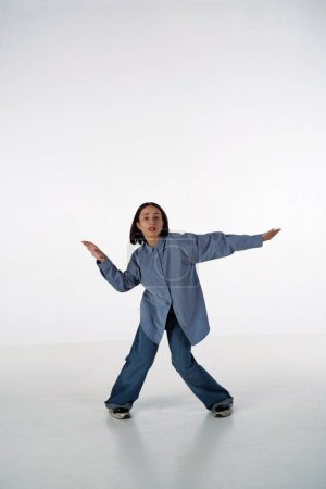 Foto de Una joven en ropa de mezclilla casual posa en un estudio sobre un fondo blanco. La bailarina demuestra los elementos coreográficos de una danza experimental de estilo hip hop - Imagen libre de derechos