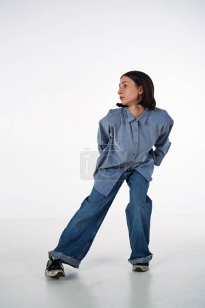 Photo for A young woman in denim casual wear poses in a studio against a white background. The dancer demonstrates the choreography elements of an experimental hip hop style dance - Royalty Free Image