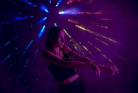 Photo for A young woman in black casual clothes poses in a studio with smoke, purple light and beams of multicolored light. The dancer demonstrates elements of experimental hip hop style dance choreography - Royalty Free Image