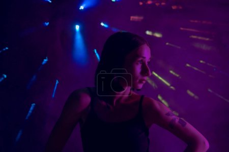 Photo for A young woman in black casual clothes poses in a studio with smoke, purple light and beams of multicolored light. The dancer demonstrates elements of experimental hip hop style dance choreography - Royalty Free Image