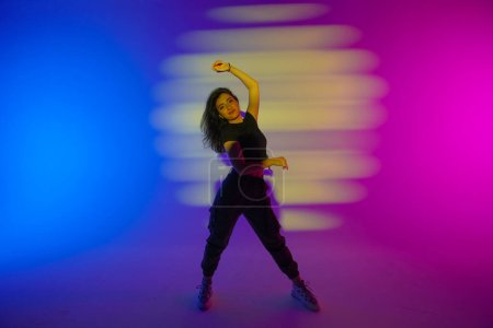 Young woman in black clothes posing in studio with gradient blue, yellow, pink neon light. Dancer demonstrates elements of jazz funk style dance. Contemporary choreography