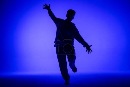 Téléchargez les photos : Silhouette de mec en tenue décontractée dansant des éléments de hip hop en studio avec lumière bleue. Le danseur fait preuve de plasticité corporelle. Pleine hauteur. chorégraphie de rue moderne - en image libre de droit