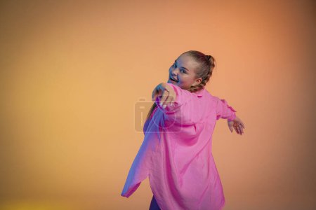 Téléchargez les photos : Une jeune femme en chemise rose pose sur un fond de studio orange. La danseuse montre des éléments de chorégraphie jazz-funk. chorégraphie moderne - en image libre de droit
