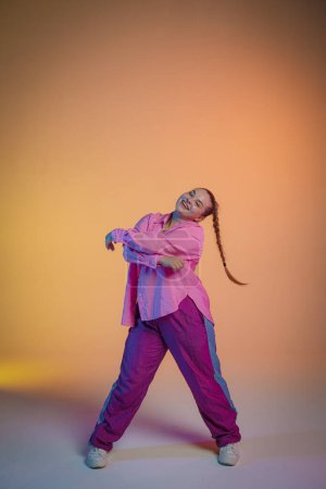 Jovencita enérgica vestida de rosa bailando a los ritmos del jazz funk sobre el fondo de un estudio con luz naranja. La foto es perfecta para los conceptos de libertad de expresión