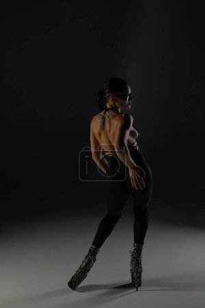 Photo for Young woman in black open clothes poses on black background of backlit studio. The dancer demonstrates elements of dance choreography in high heels. The concept of the dance promo shot - Royalty Free Image