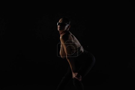Téléchargez les photos : Jeune femme en corsage noir et collants pose sur fond studio noir rétroéclairé. Une danseuse montre des éléments de danse sur des talons hauts. Danse promo concept vidéo - en image libre de droit