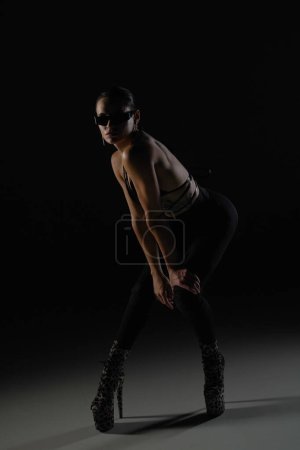 Foto de Mujer joven en corpiño negro y medias posa sobre fondo negro estudio retroiluminado. Bailarina femenina demuestra elementos de danza en tacones altos. Danza concepto de vídeo promocional - Imagen libre de derechos