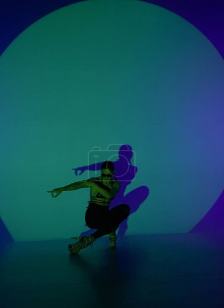 Téléchargez les photos : Une jeune femme pose dans un studio sombre avec la lumière bleue comme centre de projecteurs. L'ombre de son corps se reflète sur le mur. Une danseuse montre des éléments de danse en talons hauts - en image libre de droit