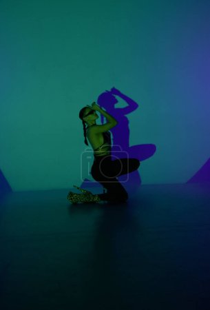 Foto de Una joven posa en un estudio oscuro con luz azul como centro de atención. La sombra de su cuerpo se refleja en la pared. Una bailarina muestra elementos de danza en tacones altos - Imagen libre de derechos