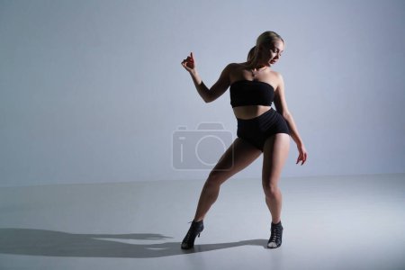 Photo for Female dancer in black shorts and top dancing in high heels. Young woman posing and showing body flexibility in studio on white background - Royalty Free Image