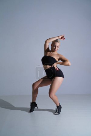 Foto de Bailarina femenina en pantalones cortos negros y top bailando en tacones altos. Mujer joven posando y mostrando flexibilidad corporal en estudio sobre fondo blanco - Imagen libre de derechos
