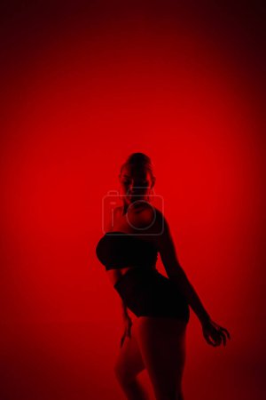 Téléchargez les photos : Silhouette danseuse en short noir et top dansant sur talons hauts. Jeune femme pose gracieusement et montre mince corps flexible en studio sombre avec lumière rouge - en image libre de droit