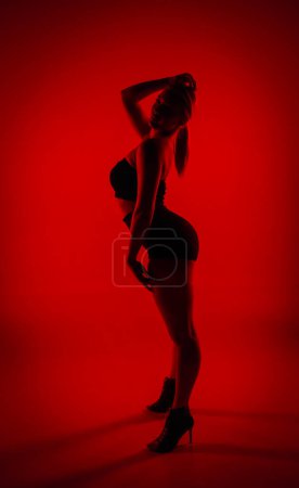Foto de Bailarina femenina Silhouette en pantalones cortos negros y top bailando en tacones altos. Mujer joven posa con gracia y muestra cuerpo delgado y flexible en estudio oscuro con luz roja - Imagen libre de derechos