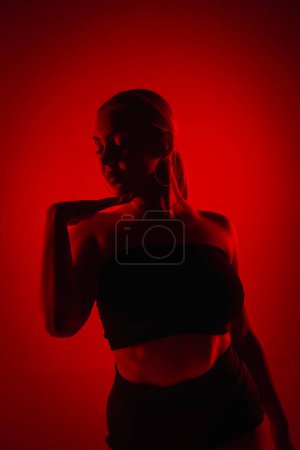 Photo for Silhouette female dancer in black shorts and top dancing on high heels. Young woman poses gracefully and shows off slender flexible body in dark studio with red light - Royalty Free Image