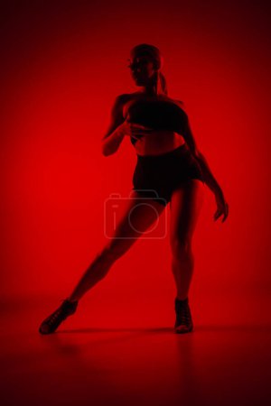 Foto de Bailarina femenina Silhouette en pantalones cortos negros y top. Mujer joven posa con gracia y muestra cuerpo delgado y flexible en estudio oscuro con luz roja. Elementos de la coreografía de baile de tacón alto - Imagen libre de derechos
