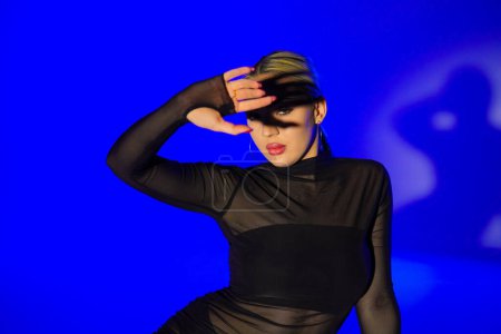 Photo for Female dancer in black tight fitting clothes posing, shadow from her hand falls on her face. Young woman showing off slender flexible body in dark studio with blue light. Elements of high heels dance - Royalty Free Image